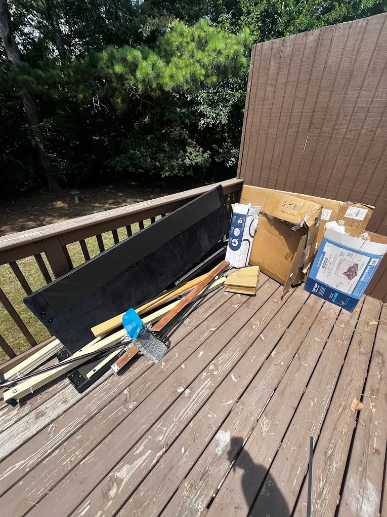 A raised back deck stacked with flattened cardboard boxes, lengths of timber, a headboard and garden tools leaning against the railing
